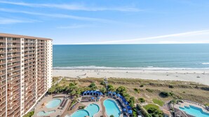 Un hotel moderno con numerosi balconi che si affaccia su una spiaggia e su una zona piscina con ombrelloni.