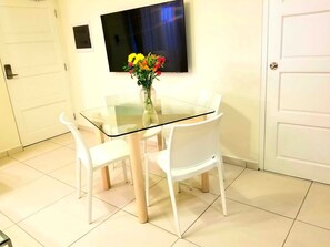 Une table à manger avec un plateau en verre, des chaises blanches, un vase rempli de fleurs et un téléviseur à écran plat fixé au mur.