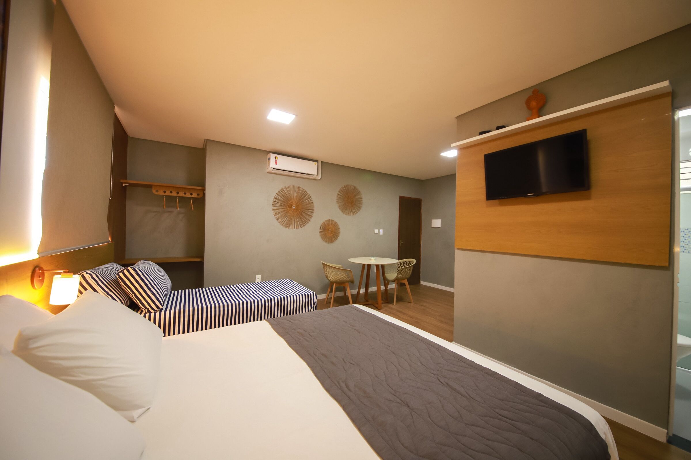 A hotel room with a bed, bedside table, lamp, wall-mounted TV, and a small dining area with chairs and a table.