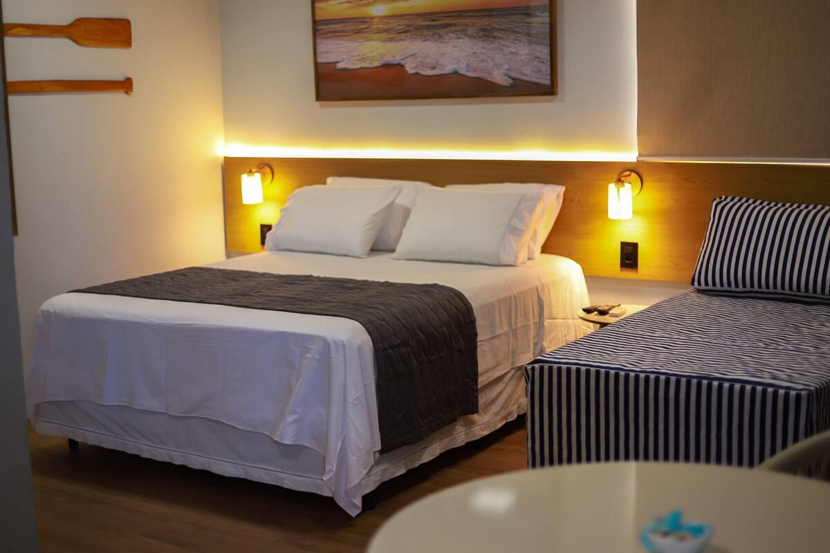Ein Hotelzimmer mit einem großen Bett, Nachttischlampen, einem kleinen Tisch und einem Bild mit einer Strandansicht.