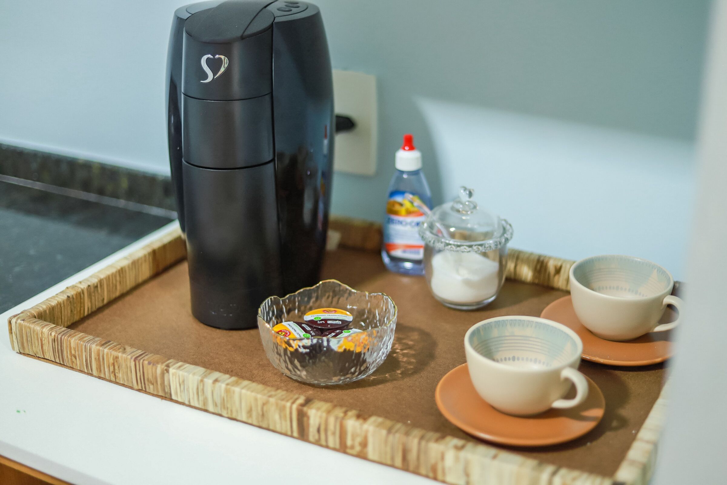 Uma bandeja com uma cafeteira, uma tigela de vidro com acompanhamentos e duas xícaras sobre uma mesa.