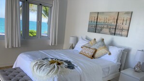 Quarto com uma cama grande, um banquinho, uma mesinha de cabeceira e uma pintura de uma cena de praia.