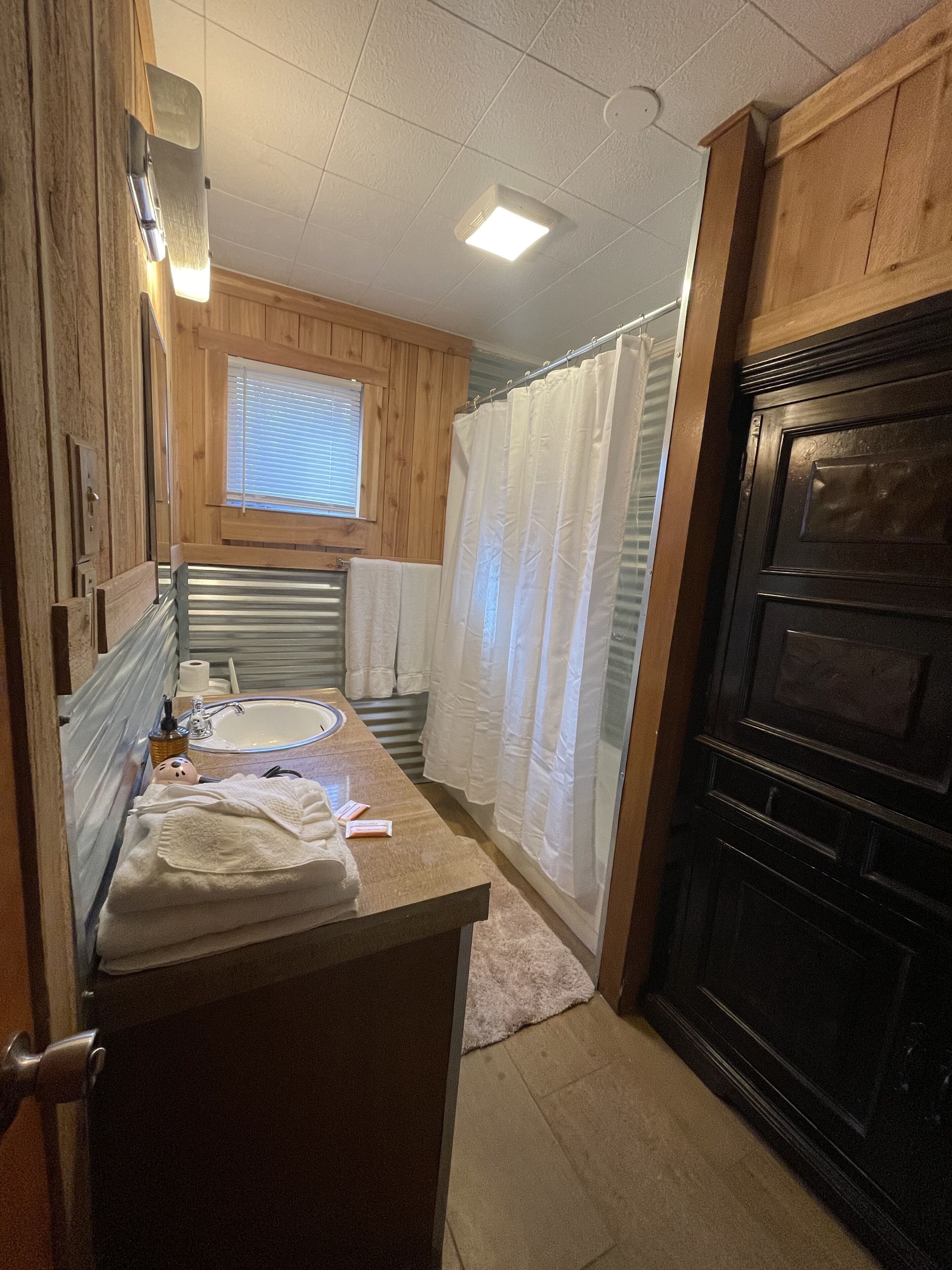 A compact bathroom with a wooden vanity, a sink, a window with blinds, a shower with a white curtain, and a dark wooden door.