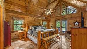 A wooden cabin bedroom with a bed, bench, and a ceiling fan. - Highland Nook - Pet Friendly | Mountain View | Hot Tub | 3 Kings | Finnish Sauna (Morganton)