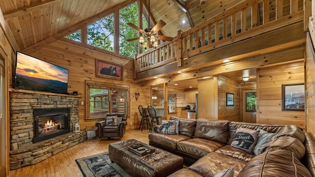 A cabin interior with a stone fireplace, leather couches, a dining area, and a balcony with a wooden railing. - Highland Nook - Pet Friendly | Mountain View | Hot Tub | 3 Kings | Finnish Sauna (Morganton)
