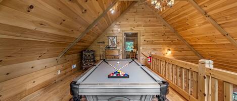 A pool table in a wooden room with a sloped ceiling and a ceiling fan.