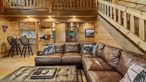A wooden cabin interior with a large leather sofa, a dining area with two chairs, and a staircase leading to an upper level. - Highland Nook - Pet Friendly | Mountain View | Hot Tub | 3 Kings | Finnish Sauna (Morganton)