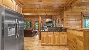 A wooden cabin interior with a kitchen featuring a stainless steel refrigerator, wooden cabinets, and a granite countertop. - Highland Nook - Pet Friendly | Mountain View | Hot Tub | 3 Kings | Finnish Sauna (Morganton)