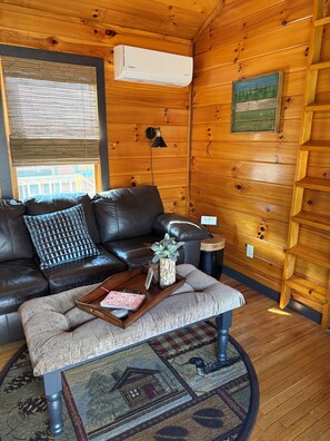 Ein gemütliches Blockhaus-Interieur mit einem Ledersofa, einem Holztisch, einem runden Teppich und einer Wand-Klimaanlage.