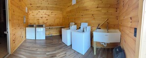 A compact room with wooden walls and a wooden floor, containing two washing machines, a sink, and a shower area.