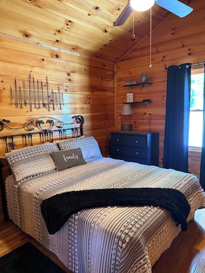 A wooden cabin bedroom with a bed, a nightstand, a wall-mounted clock, and a ceiling fan.