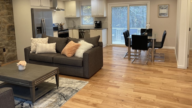 A modern living room with a grey sofa, coffee table, and a dining area with a black table and chairs. A kitchen with white cabinets and a stone wall is visible in the background. - Home w/game room hot tub 65" TV sauna AC (Albrightsville)