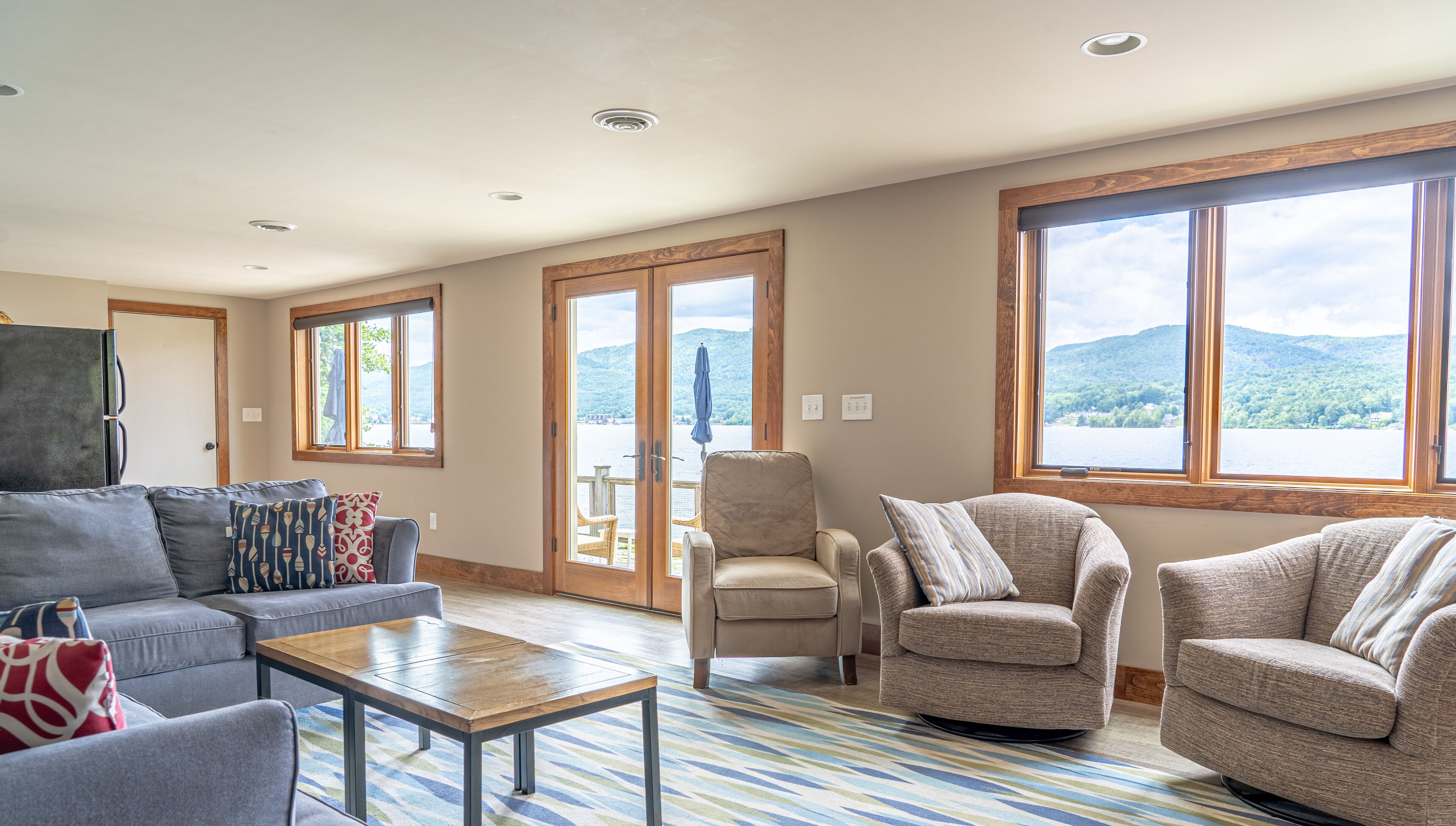 A spacious living room with a large window offering a lake view, a grey sofa, a beige armchair, and a wooden coffee table.