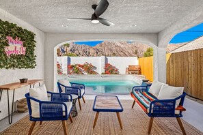 Zona junto a la piscina con sillas de mimbre azules, una mesita y un ventilador de techo.