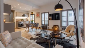 Un salon moderne avec un canapé, une table basse et un coin repas comprenant des chaises et une table. On aperçoit une cuisine à l’arrière-plan, avec une cuisinière et des placards.