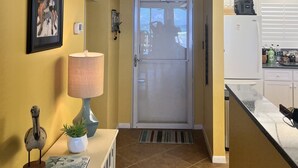 En hall med ett träpanelat tak, en inramad tavla på väggen, en lampa, ett litet bord med ett anteckningsblock, en krukväxt och en dörr som leder till ett köksområde.