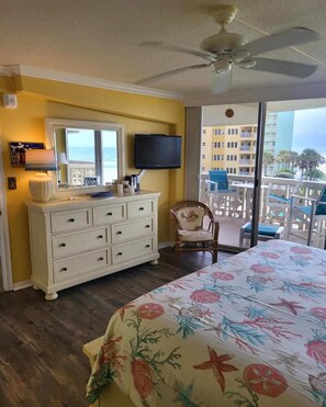 A hotel room with a bed, a dresser, a television, a chair, and a balcony with a view of the ocean and buildings.