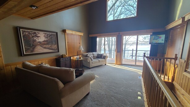 A spacious living room with a high ceiling, a large window, a sofa, a television, and a view of a deck and lake. - Lake home pontoon included.  (Clear Lake)
