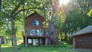 A wooden cabin with a deck and a garage, surrounded by trees and grass. - Lake home pontoon included.  (Clear Lake)