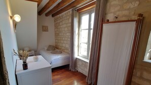A small bedroom with a single bed, a white desk, a window with curtains, and a stone wall.