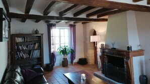 A cozy living room with exposed wooden beams, a fireplace, a bookshelf, a leather sofa, and a large window with curtains.