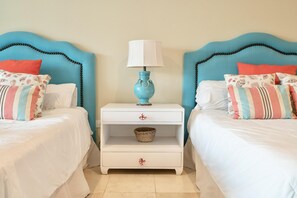 A room with two beds, each with a blue headboard and white bedding. There is a white nightstand with a blue lamp and a woven basket between the beds. - Beachfront Lux condo w/ private terrace (Cap Cana)
