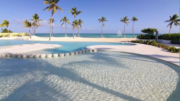 Piscina de resort com água cristalina, rodeada por palmeiras e uma praia.