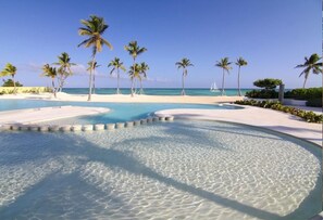 A resort pool with clear water, surrounded by palm trees and a beach. - Beachfront Lux condo w/ private terrace (Cap Cana)