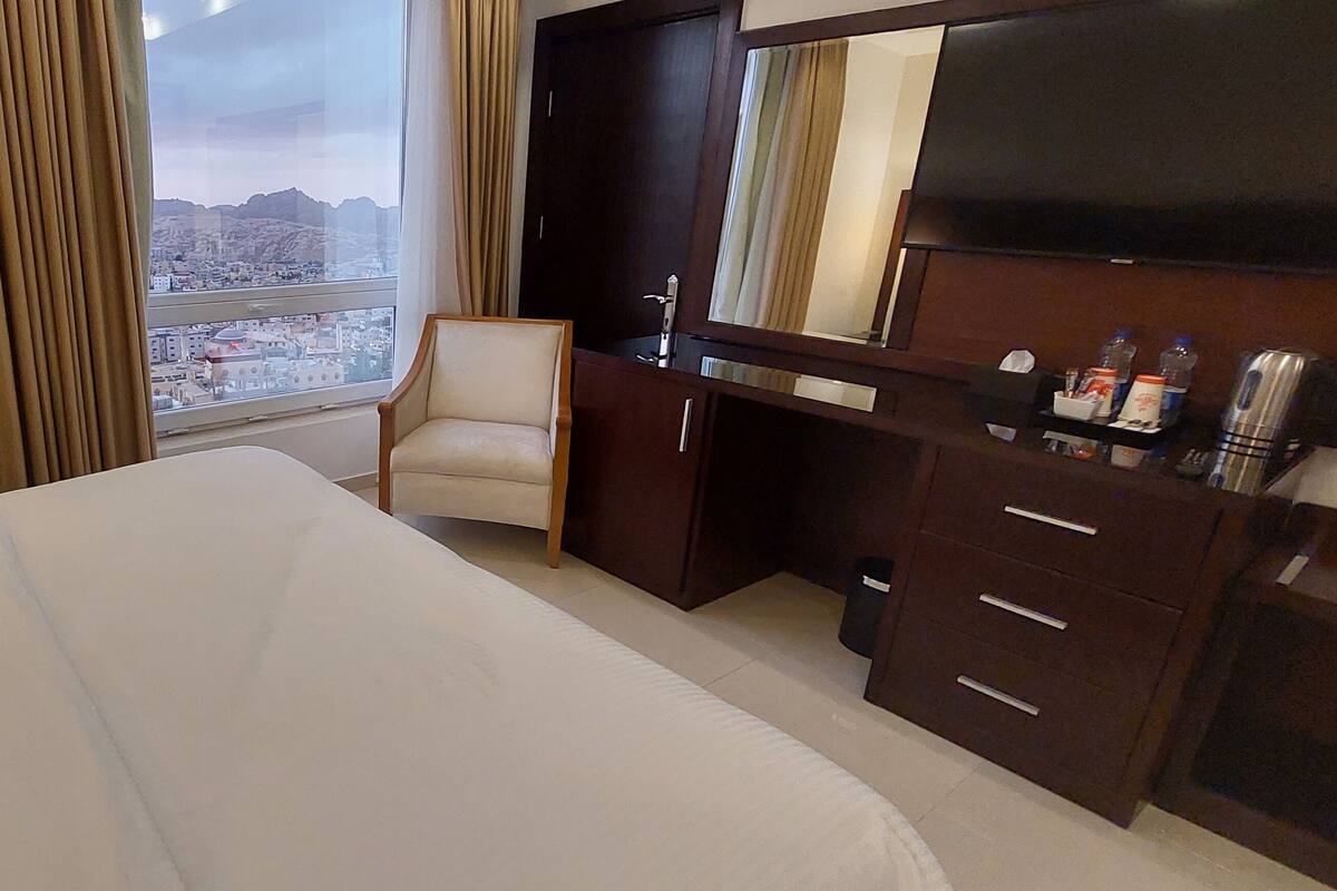 A hotel room with a large bed, a desk with a chair, a mirror, and a view of the cityscape.