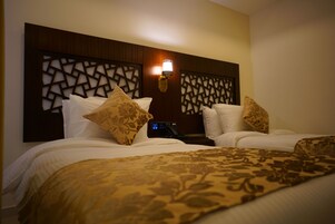 Habitación de hotel con dos camas, cada una con cubrecama y almohadas estampadas. Hay una lámpara de pared sobre las camas y un panel de control en la pared.