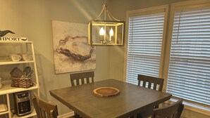 A dining area with a wooden table, four chairs, a coffee maker, a basket of kitchen utensils, and a wall-mounted light fixture.
