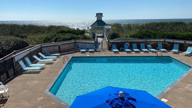A rectangular pool with a wooden deck, surrounded by lounge chairs and a blue umbrella.
