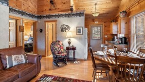 A cozy cabin interior with a wooden ceiling, a dining area with a table and chairs, a sofa, and a chair with a floral pattern.