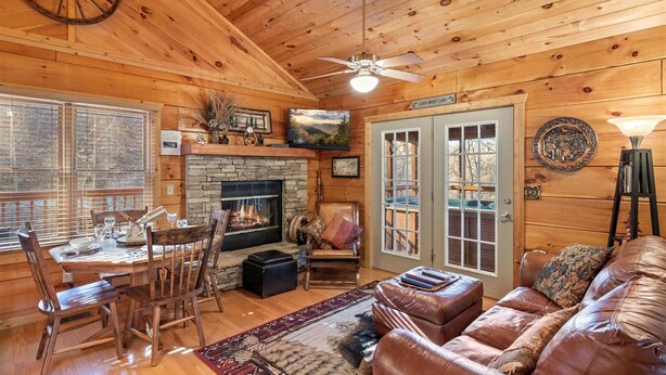 A cozy cabin interior with a stone fireplace, wooden dining table, leather sofa, and a ceiling fan.