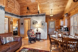 A cozy cabin interior with a wooden ceiling, a dining area with a table and chairs, a sofa, and a chair with a floral pattern.