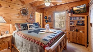 A cozy wooden cabin bedroom with a bed, a nightstand, a bookshelf, and a ceiling fan.