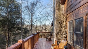 A wooden deck with a rocking chair and a table with teddy bears, overlooking a forested area.