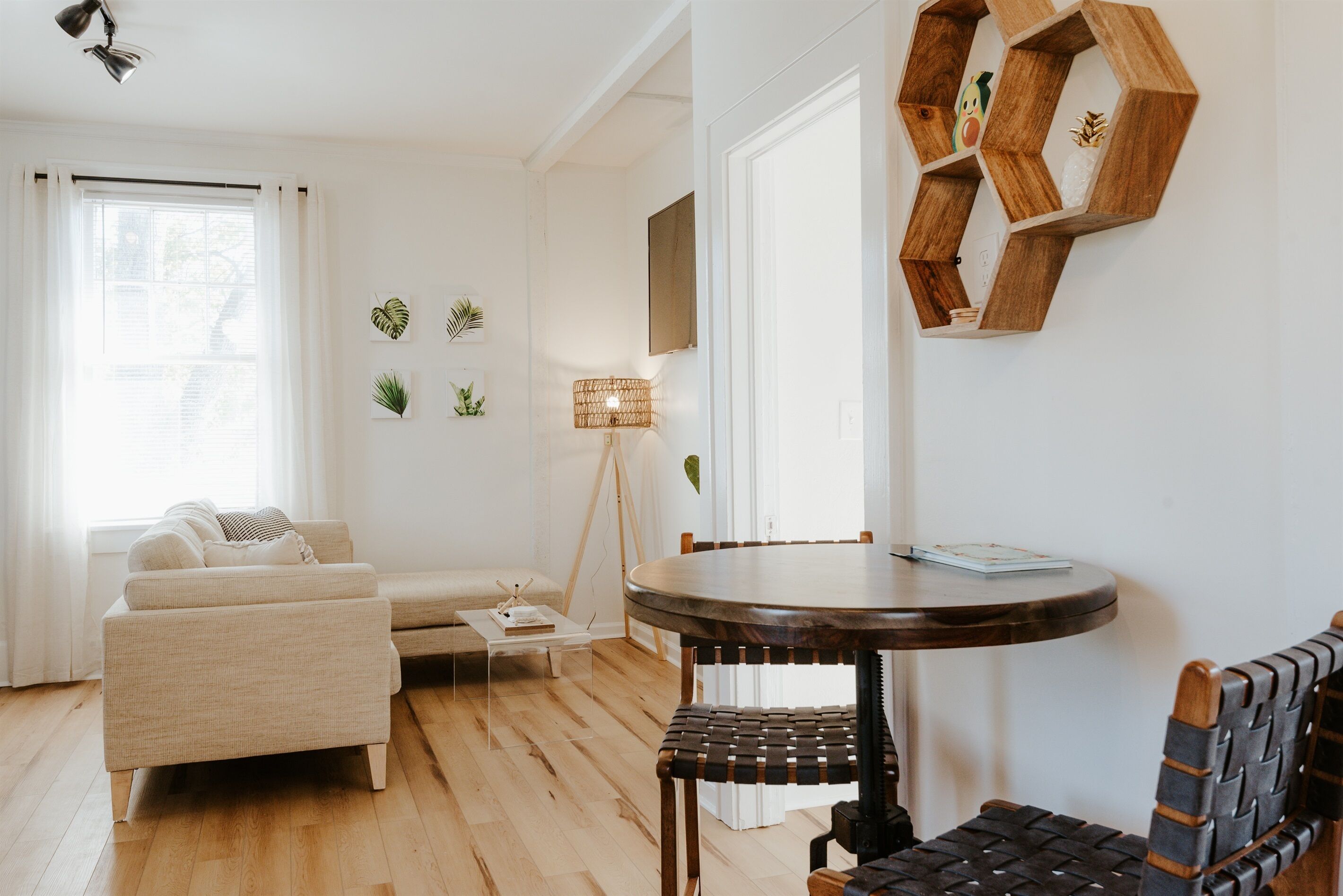 Una sala de estar con un sofá, una mesa de centro, una mesa redonda y una silla. Hay un estante hexagonal de madera en la pared y una lámpara de pie.