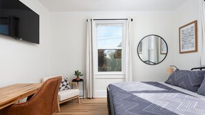 A bedroom with a bed, a desk, a chair, a window, a ceiling fan, and a mirror.