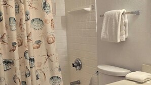 Un baño con cortina de ducha decorada con conchas marinas y estrellas de mar, un inodoro, un tocador de madera con lavamanos y un perchero para toallas.