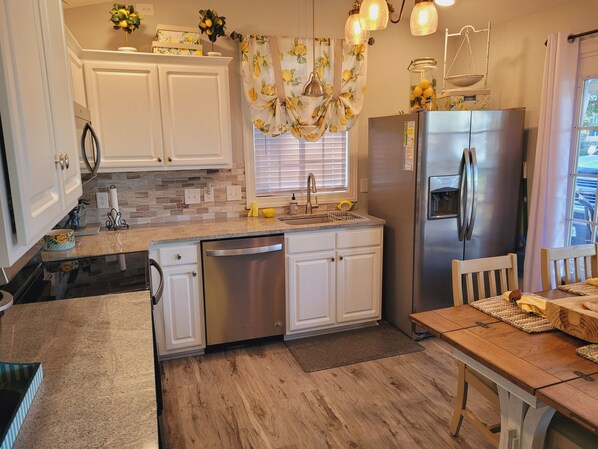A modern kitchen with white cabinets, a stainless steel dishwasher, and a refrigerator. There is a wooden dining table with chairs and a window with blinds. - 5-Star Lakefront Beach House in Ocean Lakes (Myrtle Beach)