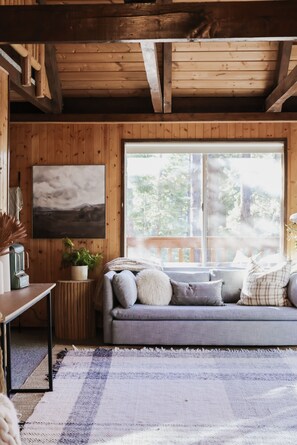 A cozy cabin interior with a wooden ceiling, a gray sofa with cushions, a large window, and a painting on the wall.