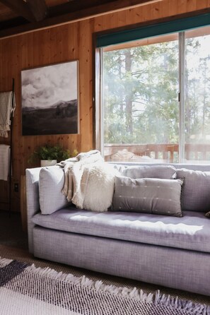 A room with a wooden wall, a grey sofa, a large window, and a framed picture.