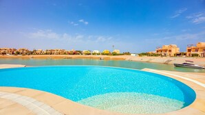 A swimming pool with clear blue water, surrounded by a tiled deck. In the background, there are buildings with a uniform architectural style. - VESTA - Luxury Villa - 5BR - Sabina (Hurghada)