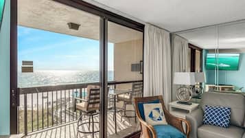 A balcony with a view of the ocean, a wicker chair with a cushion, a grey sofa, and a table with a lamp.