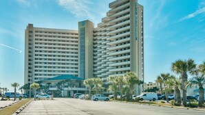 A modern hotel with a central atrium, multiple floors, and a parking area with cars and palm trees.