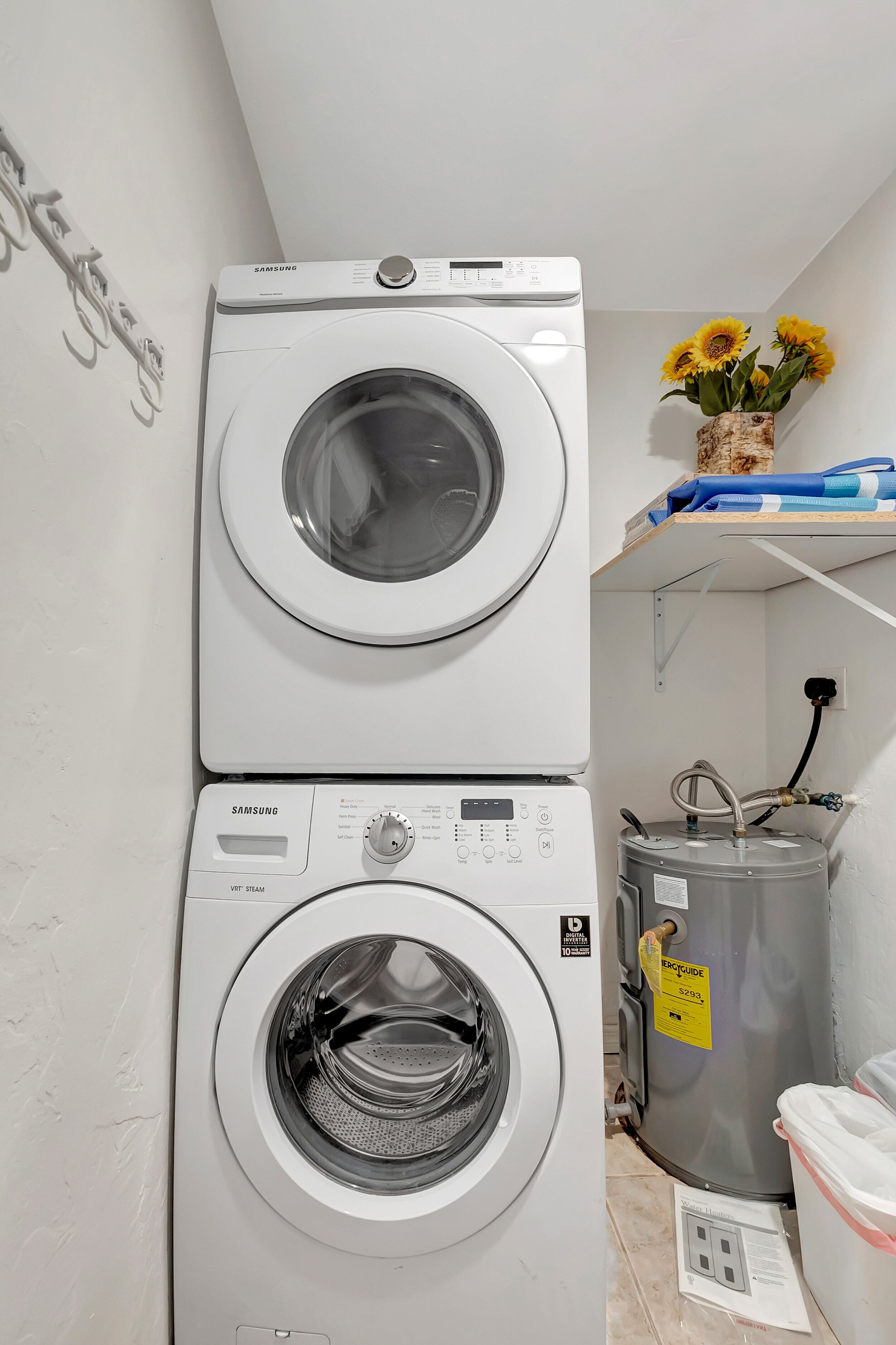 A stacked washer and dryer unit with a water heater and a container on the floor.