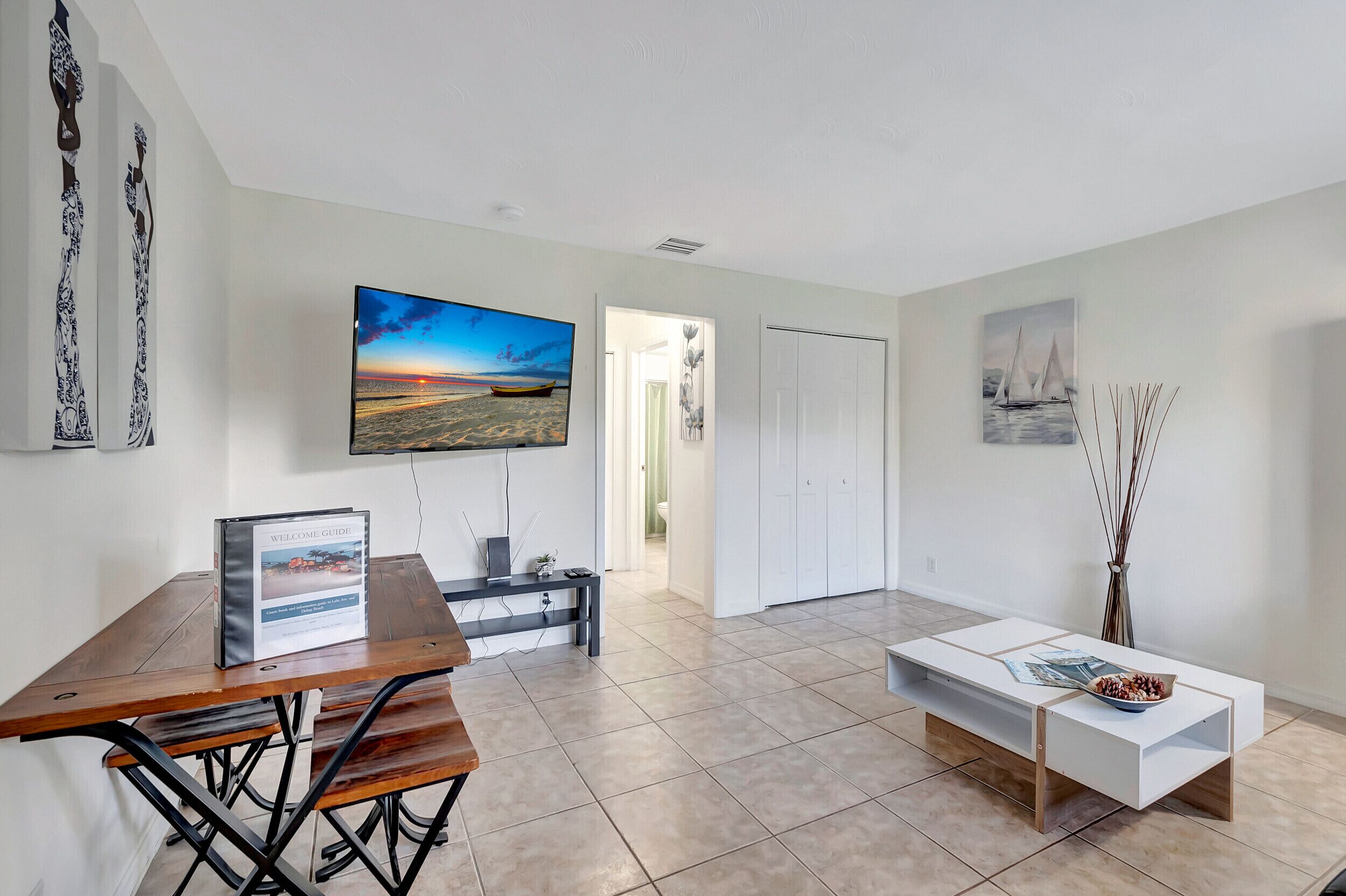 A modern living room with a wooden dining table, a white sofa, and a flat-screen TV mounted on the wall.
