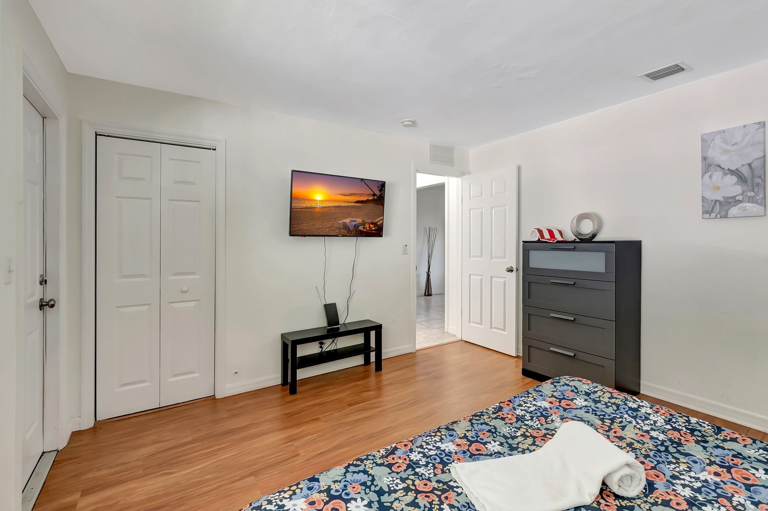 Un dormitorio con cama, televisor montado en la pared, cómoda y puerta que da a otra habitación.