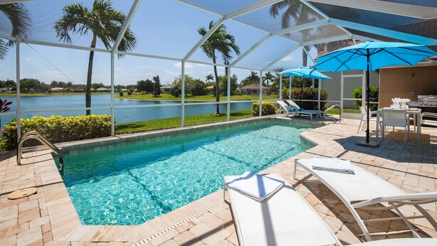 Une piscine avec vue sur un lac, entourée de palmiers et dotée d’un patio couvert avec des chaises longues et des parasols.